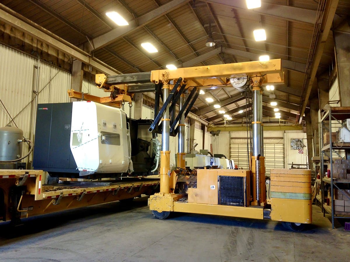 A CNC machine being lifted onto a flatbed trailer with a hydraulic gantry during a machine move
